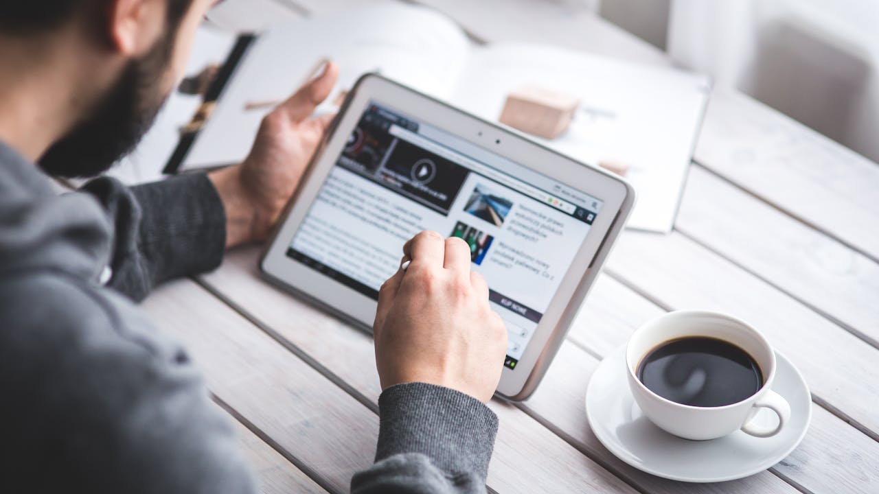 A man uses a tablet to browse news while enjoying a cup of coffee, stylus in hand.