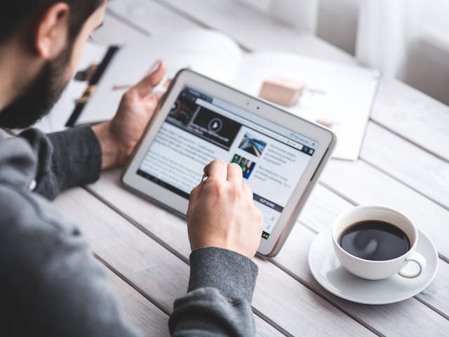 A man uses a tablet to browse news while enjoying a cup of coffee, stylus in hand.
